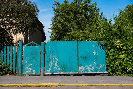 An old iron green gate with cracked paint near a residential building with a garden.