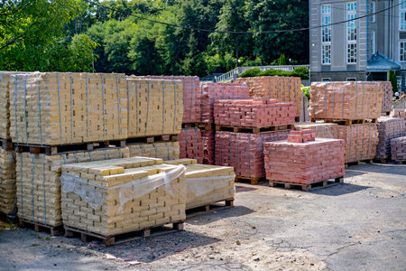 A large pile of bricks in various colors stacked on wooden pallets in a lot with a building in the background.の写真素材