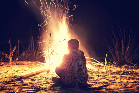 Young boy sit near a bright bonfire