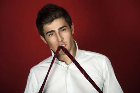 Beautiful  young man in a white shirt holds a red tie with his teethの写真素材