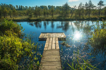 Wooden footbridge on a small lake with a bright sun glare on the water.の写真素材