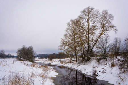 Landscape with a small river in snow-covered shores in a winter overcast day.の写真素材