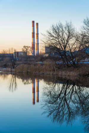 Beautiful evening cityscape with a mirror image of trees and tall pipes in a river.の写真素材
