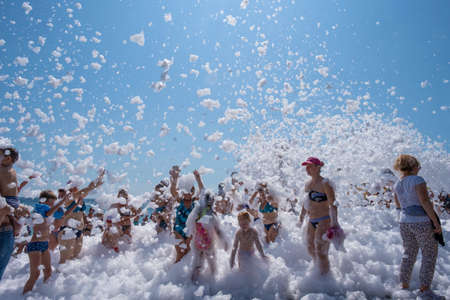 Yalta, Crimea - 07/09/2019: Flakes of snow-white foam fall asleep to cheerful people on the beach on a sunny day.のeditorial素材