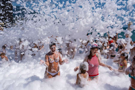 Yalta, Crimea - 07/09/2019: Flakes of snow-white foam fall asleep to cheerful people on the beach on a sunny day.のeditorial素材