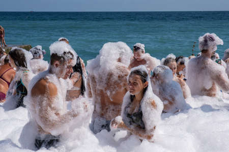 Yalta, Crimea - 09/07/2019: Flakes of white foam fall asleep to cheerful people on the beach on a sunny day.のeditorial素材