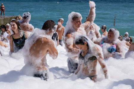 Yalta, Crimea - 09/07/2019: Flakes of white foam fall asleep to cheerful people on the beach on a sunny day.のeditorial素材