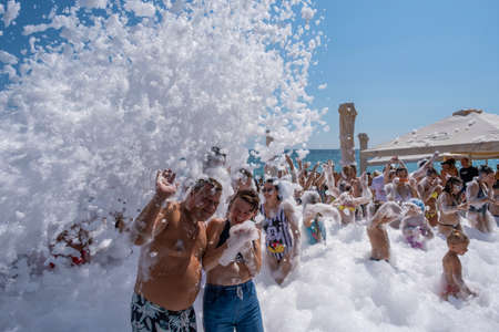 Yalta, Crimea - 09/07/2019: Flakes of snow-white foam fall asleep to cheerful people on the beach on a sunny day.のeditorial素材