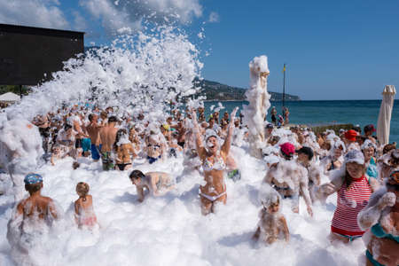 Yalta, Crimea - 07/09/2019: Flakes of snow-white foam fall asleep to cheerful people on the beach on a sunny day.のeditorial素材