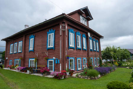 An old wooden two-story house with beautiful flower beds of various colors on a cloudy day. Vichuga...