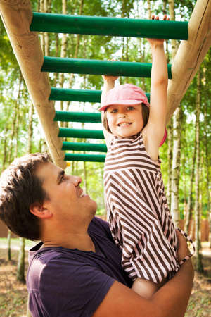 Father helping her daughter hanging on on the monkey bars in a playground, smiling and spending time together.の写真素材