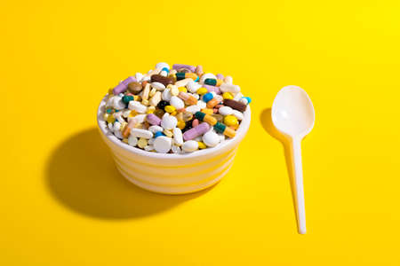 Bowl full of colorful pills on yellow background with plastic spoon. Creative composition.の写真素材