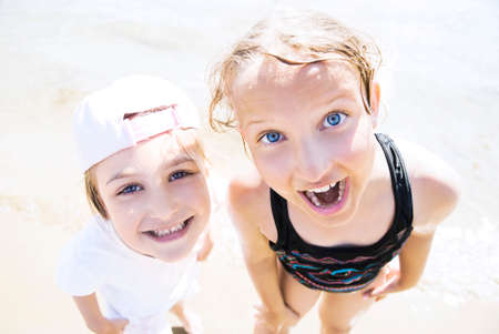 Close up sunny portrait of a two smiling sisters looking up at the camera standing on sandy beach.の写真素材