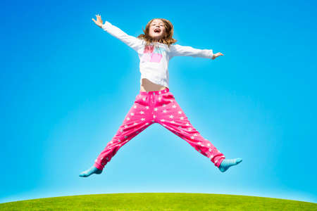 Happy girl jumping high in air over green grass against blue sky during sunny day.