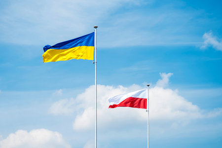 State flags of Ukraine and Poland on flagpoles over blue cloudy sky background.