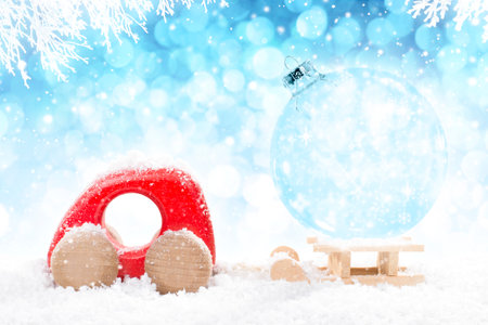 Red Wooden Car Pulling Sled with A Transparent Christmas Ball in perspective on bright blue bokeh winter background with white snowy branches and falling snow.の写真素材