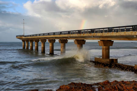 Pier jutting out into the stormy sea and a small rainbow in the cloudsの写真素材