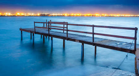 Long exposure of an old jetty on a river with city lights in the backgroundの写真素材