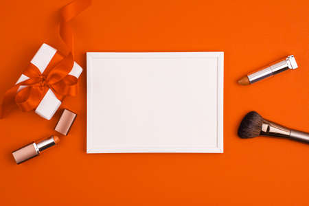 Mockup white frame white gift present with red bow, make up brushes and lipstick on vibrant orange background. Cosmetics and beauty concept. Make up concept with copy space template flat layの写真素材
