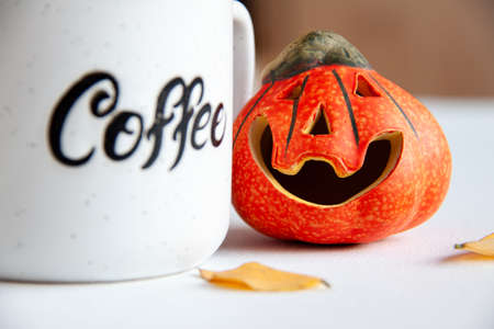 Ceramic orange small smile halloween pumpkin with autumn leaves near it and coffee cup with text "coffee" on it. Holiday conceptの写真素材