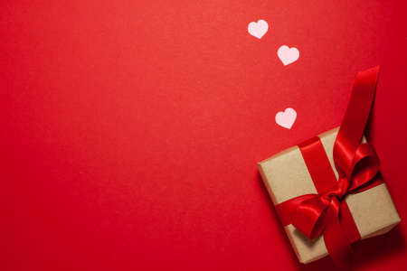 Small pink hearts near the brown paper gift box with a red ribbon bow. Valentines day concept. Flat lay top view with space for text on red backgroundの写真素材