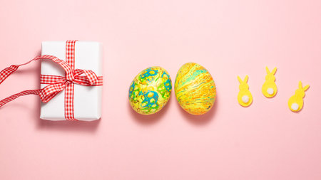Easter eggs, white gift box with red gift bow and yellow rabbits on pink background. Flat lay top...