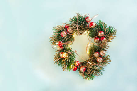 Christmas round wreath from fake green tree branches and red berries which painted in gold at the tips, Christmas lights that wrap a wreath on bright blue background with space for textの写真素材