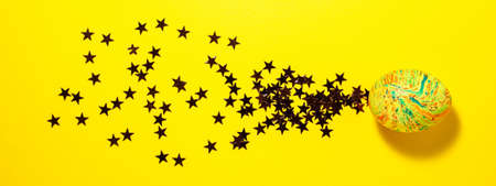 Easter colorful egg flies through the frame leaving stars behind itself on yellow background. Flat lay bannerの写真素材