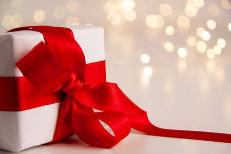 Close up of white paper gift box with a red satin ribbon bow on bright background with golden bokeh. Holidays greeting cards and Christmas shopping concept with copy space on the right.の写真素材