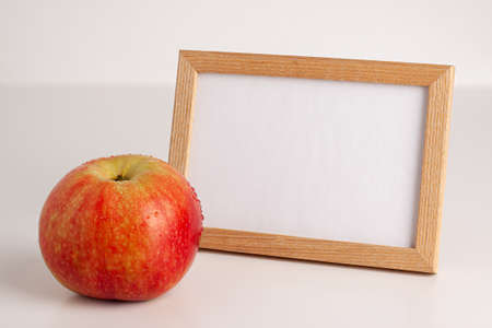 Close up mockup of red yellow apple with water drops and white frame with wooden borders on bright background. Healthy organic food, autumn mood and still life concept with copy spaceの写真素材