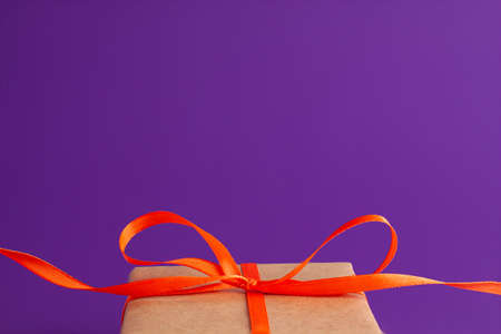 Close up of brown gift box with a orange satin ribbon bow on the bottom on vibrant purple blurred background with copy space. Holiday autumn and Halloween conceptの写真素材