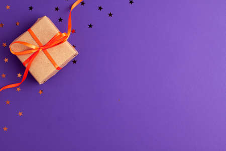 Flat lay of brown gift box with a orange satin ribbon bow and stars shapes on vibrant purple...