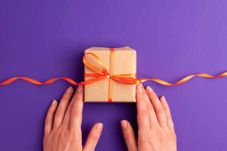 Flat lay of brown gift box with a orange satin ribbon bow and hands on it on vibrant purple background with copy space. Holiday autumn and Halloween conceptの写真素材