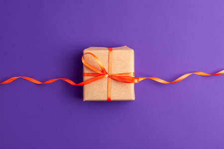 Flat lay of brown gift box with a orange satin ribbon bow on vibrant purple background with copy space. Holiday autumn and Halloween concept.の写真素材