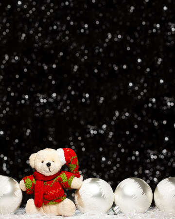 Christmas decorations view of teddy bear in red green sweater and Christmas hat and four silver evening balls with white snow on it on dark background with silver colors bokeh. Holidays conceptの写真素材