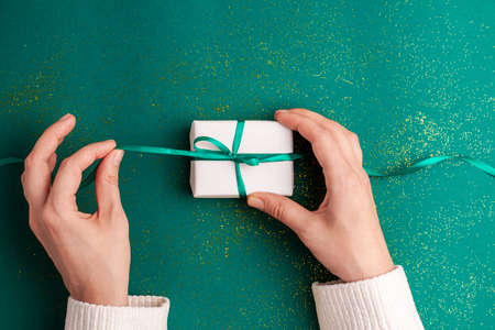 Human hands in white sweater untied white gift box with a emerald green satin ribbon bow on green...