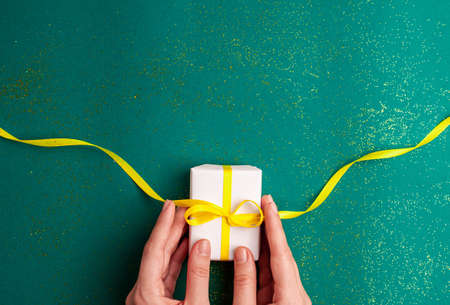 Human hands holding white gift box with a yellow satin ribbon bow on emerald green background with...
