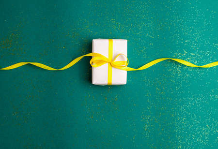 White gift box with a yellow satin ribbon bow on emerald green background with gold colored glitter....