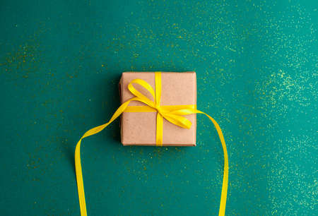 Brown gift box with a yellow satin ribbon bow on emerald green background with gold colored glitter. Christmas Holidays and present concept flat layの写真素材