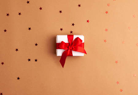 White gift box with a red satin ribbon bow tied on brown background with red stars shapes.Christmas Holidays and present concept flat lay.の写真素材
