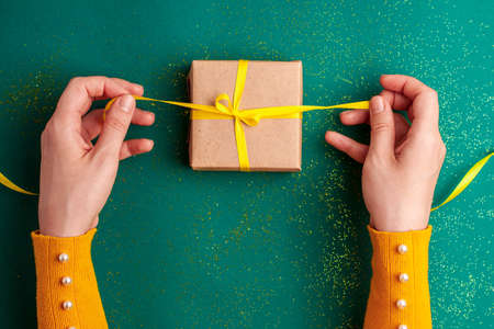 Human hands untied brown gift box with a yellow satin ribbon bow on emerald green background with...