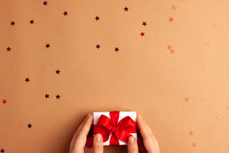 Human hands puts a white paper gift with a red satin ribbon bow on brown background with red stars shapes. Christmas Holidays concept flat lay with copy spaceの写真素材