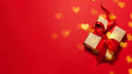 Brown paper gift box with a red satin ribbon bow on red background with gold bokeh of heart shapes on the right with copy space. Flat lay Valentines day father mother day and presents conceptの写真素材
