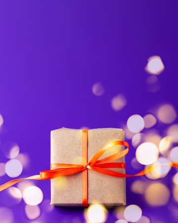 Close up of brown gift box with an orange satin ribbon bow at the bottom and golden bokeh on vibrant purple blurred background with copy space. Holiday presents and Halloween concept.の写真素材