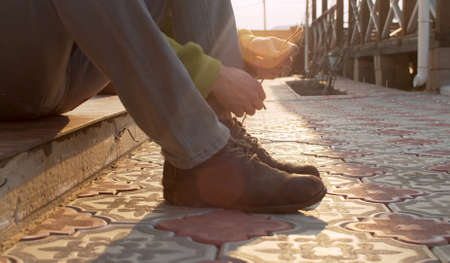 Close up male hands tying shoelaces on boots. A young man sitting in the tiled courtyard of a country house. Lens flareの写真素材