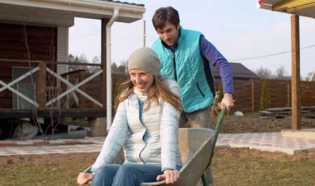 Young man pushing his wife in a wheelbarrow. Happy family playing in a spring garden. Both laughing. Front viewの写真素材
