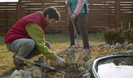 Family prepares flowerbed for spring planting. Son weeding flowerbed, mom raking dry grass with rakeの写真素材