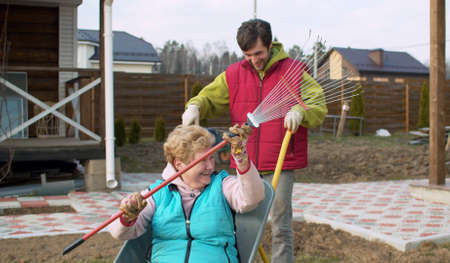 A young man riding his elderly mother in a wheelbarrow. Mom with a rake in her hands sits in a wheelbarrow and laughsの写真素材