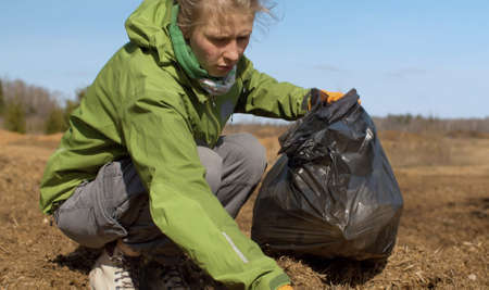 Close up woman eco volunteer picking trash a in black garbage bag on the forest glade. Early spring, bright blue sky, no leaves on the trees, windy weather. Earth day. Eco friendly conceptの写真素材