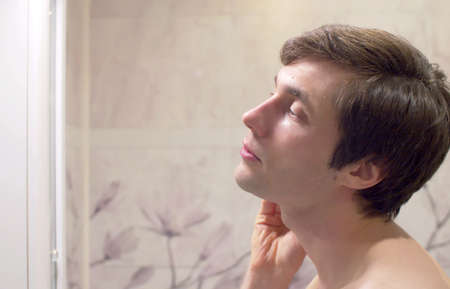 Close up portrait of young handsome man applying tonic on his face in the bright bathroom. Face skin...